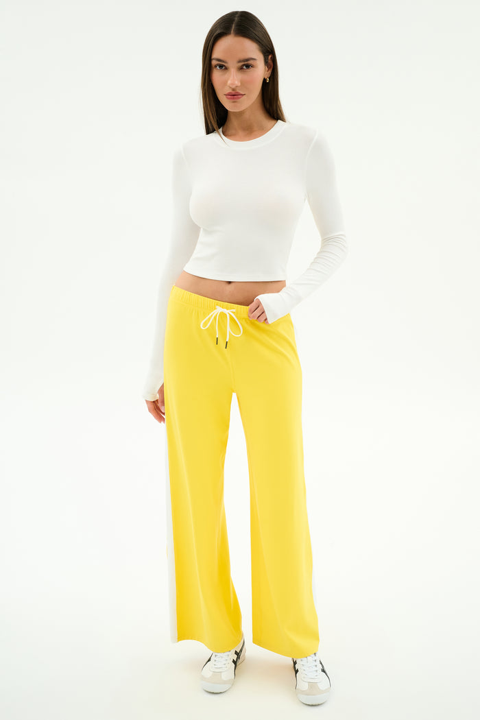 A woman poses against a white background wearing SPLITS59’s Willow Airweight Wide Leg pants in Bright Yellow/White, paired with a long-sleeve white spandex crop top and white sneakers.