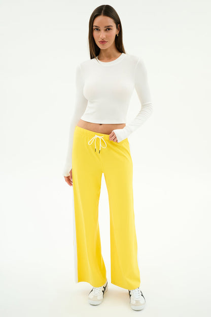 A woman poses against a white background wearing SPLITS59’s Willow Airweight Wide Leg pants in Bright Yellow/White, paired with a long-sleeve white spandex crop top and white sneakers.