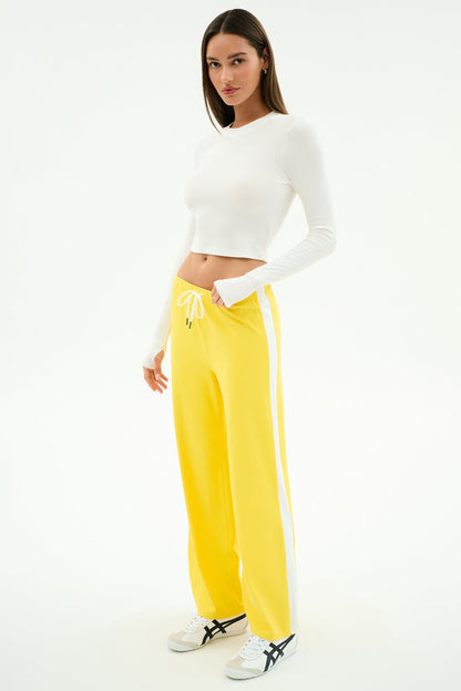 A woman stands against a white background wearing a white spandex long-sleeve crop top, SPLITS59 Willow Airweight Wide Leg pants in Bright Yellow/White, and white sneakers.