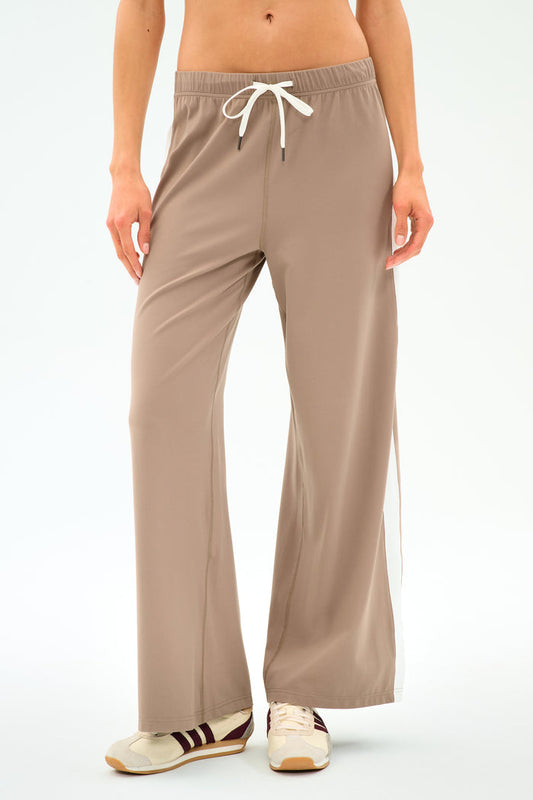 A person wears SPLITS59 Willow Airweight Wide Leg pants in Latte/White, paired with beige sneakers with brown stripes, standing against a plain white background.