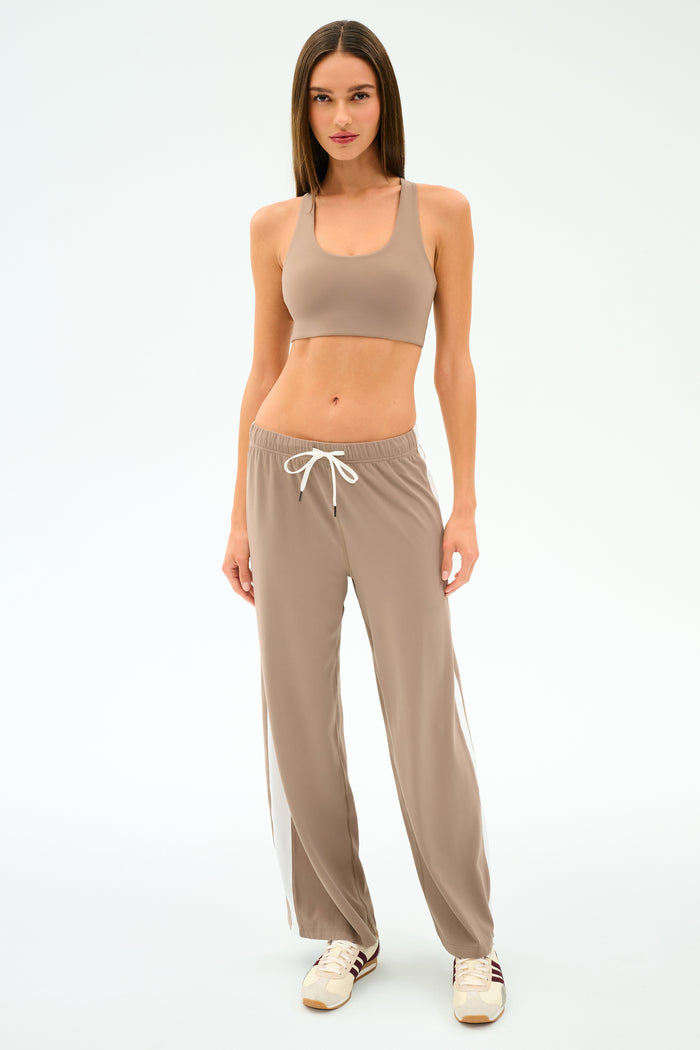 A woman faces forward in SPLITS59 Willow Airweight Wide Leg pants in Latte/White, paired with a taupe spandex sports bra and beige sneakers against a plain white background.