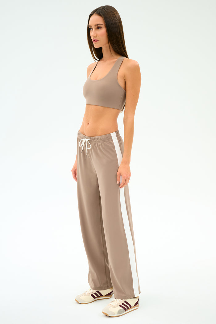 A woman stands in profile wearing SPLITS59’s Willow Airweight Wide Leg pants in Latte/White, a beige sports bra, and beige sneakers with brown stripes.